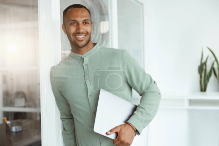 Téléchargez les photos : Homme d'affaires africain souriant tenant un ordinateur portable tout en se tenant dans un bureau confortable. Concept d'entreprise - en image libre de droit