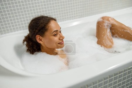 Foto de Hermosa mujer afroamericana toma baño de burbujas en la bañera y relajarse. Concepto de spa y bienestar - Imagen libre de derechos