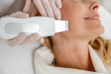 Foto de Cosmetólogo profesional está haciendo limpieza facial ultrasónica para hermosa mujer rubia madura feliz en la clínica moderna, de cerca - Imagen libre de derechos