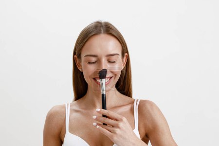 Téléchargez les photos : Jeune femme avec une peau saine maquillage à l'aide d'une brosse cosmétique sur fond de studio - en image libre de droit