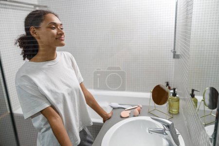 Téléchargez les photos : Femme africaine souriante debout dans la salle de bain et regardant dans le miroir. Photo de haute qualité - en image libre de droit