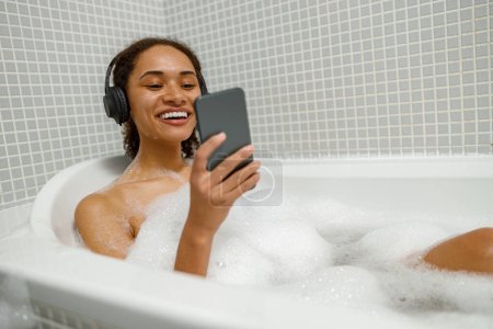 Téléchargez les photos : Afro femme américaine dans les écouteurs écouter de la musique et utiliser un smartphone lors de la prise d'un bain - en image libre de droit