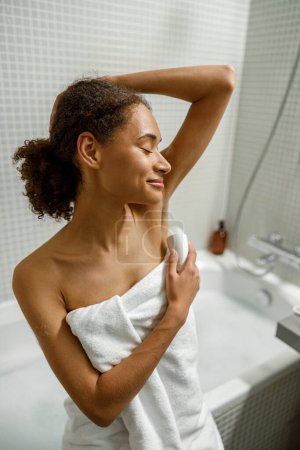 Téléchargez les photos : Afro femme américaine en serviette utilisation déodorant sous les bras bâton pour l'hygiène quotidienne assis dans la salle de bain - en image libre de droit