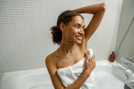 Photo for Afro american woman in towel use underarm deodorant stick for daily hygiene sitting in bathroom - Royalty Free Image