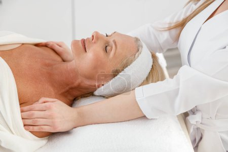 Téléchargez les photos : Vue latérale d'une belle patiente d "âge moyen détendue allongée dans une armoire de beauté lors d'une séance de massage - en image libre de droit