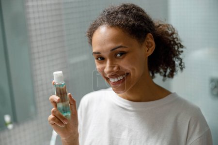 Téléchargez les photos : Femme africaine souriante tenant des cosmétiques naturels pour la peau debout dans la salle de bain. Photo de haute qualité - en image libre de droit
