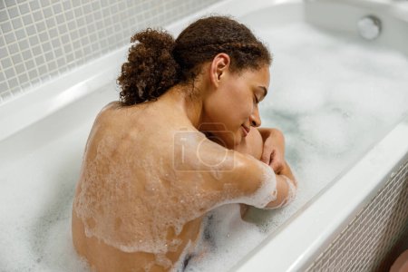 Téléchargez les photos : Belle femme afro-américaine prend un bain moussant dans la baignoire et se détendre les yeux fermés - en image libre de droit