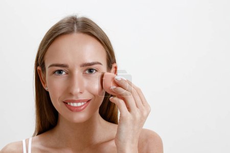 Téléchargez les photos : Jeune femme tenir éponge rose pour l'application de produits cosmétiques de beauté sur le visage. Photo de haute qualité - en image libre de droit