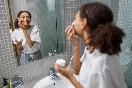 Foto de Mujer sonriente aplicando crema anti-arrugas de pie detrás del espejo en el baño casero. foto de alta calidad - Imagen libre de derechos