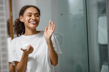 Téléchargez les photos : Femme souriante appliquant crème anti-rides debout derrière le miroir dans la salle de bain à la maison.Photo de haute qualité - en image libre de droit