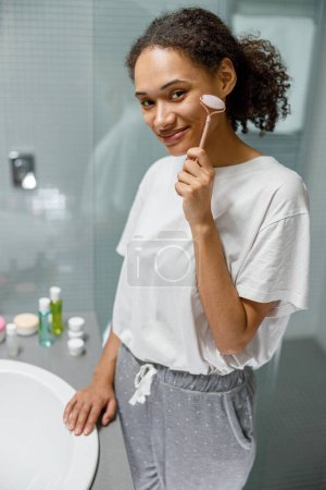 Téléchargez les photos : Femme faisant massage du visage à l'aide d'un rouleau de visage avec pierre de quartz dans la salle de bain. Photo de haute qualité - en image libre de droit
