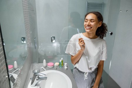 Photo for Smiling woman brushing teeth in front of her bathroom mirror. Morning routine beauty procedure - Royalty Free Image