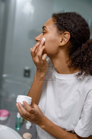Téléchargez les photos : Femme appliquant crème anti-rides debout derrière le miroir dans la salle de bain à la maison.Photo de haute qualité - en image libre de droit