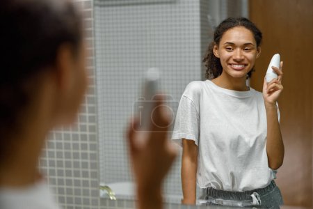 Téléchargez les photos : Femme tenant antitranspirant et souriant debout dans la salle de bain. Concept de soins corporels. Photo de haute qualité - en image libre de droit