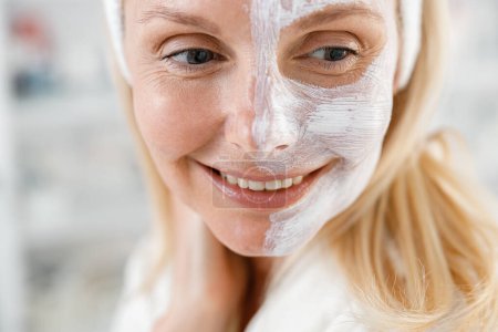 Téléchargez les photos : Gros plan de femme blonde caucasienne avec masque crème blanche sur la moitié de son visage sourit, soins de beauté - en image libre de droit