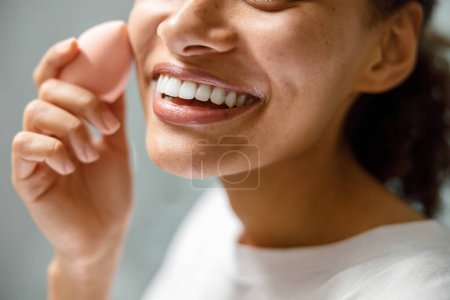 Téléchargez les photos : Gros plan de femme africaine souriante avec éponge appliquant le maquillage à la maison salle de bain. Photo de haute qualité - en image libre de droit