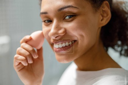 Téléchargez les photos : Gros plan de femme africaine souriante avec éponge appliquant le maquillage à la maison salle de bain. Photo de haute qualité - en image libre de droit