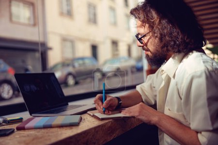 Téléchargez les photos : Étudiant mâle prenant des notes pendant la conférence en ligne tout en étant assis dans un café. Photo de haute qualité - en image libre de droit