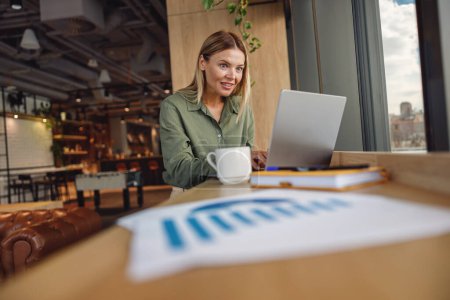 Téléchargez les photos : Femme analyste travaillant avec des documents et utiliser un ordinateur portable tout en étant assis dans le coworking près de la fenêtre - en image libre de droit