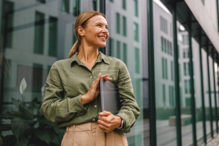 Téléchargez les photos : Femme souriante employée de bureau est debout sur le fond du bâtiment moderne et regardant côté - en image libre de droit