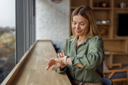 Téléchargez les photos : Femme gestionnaire souriante regardant sur sa montre-bracelet tout en étant assis dans un espace de coworking près de la fenêtre - en image libre de droit