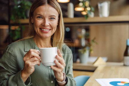 Foto de Atractiva mujer gerente sonriendo y mirando a la cámara mientras bebe café en la acogedora cafetería - Imagen libre de derechos