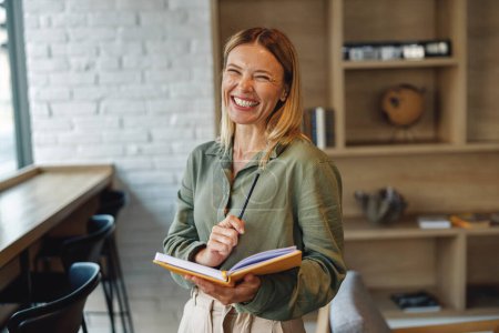 Foto de Mujer sonriente gerente toma notas en bloc de notas mientras está de pie en el fondo acogedor coworking - Imagen libre de derechos