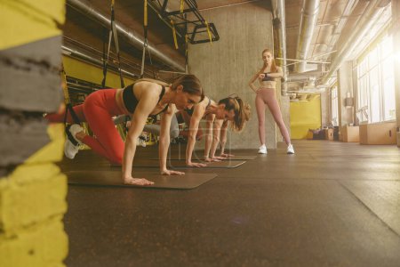 Foto de Grupo de mujeres deportistas en el gimnasio con entrenador haciendo ejercicios de entrenamiento de flexiones con TRX - Imagen libre de derechos