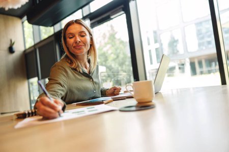 Téléchargez les photos : Femme gestionnaire souriante prenant des notes dans le bloc-notes tout en étant assis dans un coworking confortable - en image libre de droit