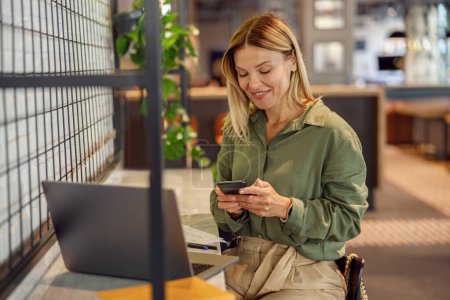 Foto de Mujer de negocios sonriente está utilizando el teléfono mientras está sentado en coworking y el trabajo en el ordenador portátil - Imagen libre de derechos