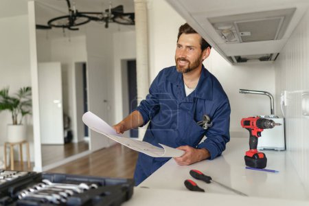 Téléchargez les photos : Homme à tout faire en uniforme debout sur la cuisine avec plan de maison et boîte à outils avant de commencer le travail - en image libre de droit