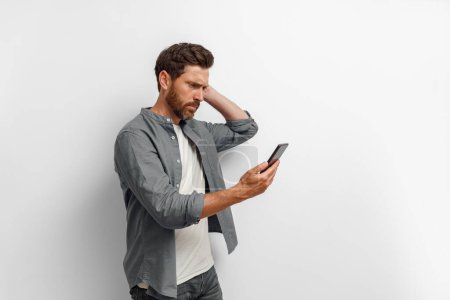 Téléchargez les photos : Homme inquiet regardant son téléphone portable tout en se tenant sur fond blanc. Photo de haute qualité - en image libre de droit