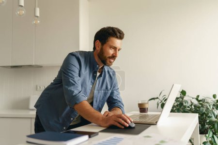 Téléchargez les photos : Homme freelance en vêtements décontractés travaillant sur ordinateur portable sur la cuisine à la maison. Concept de travail à distance - en image libre de droit