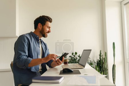 Téléchargez les photos : Homme entrepreneur tenant téléphone tout en travaillant sur ordinateur portable sur la cuisine à la maison. Concept de travail à distance - en image libre de droit