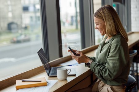 Foto de Mujer de negocios sonriente está utilizando el teléfono mientras está sentado en coworking y el trabajo en el ordenador portátil - Imagen libre de derechos