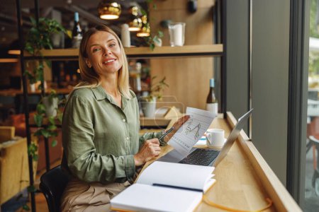 Téléchargez les photos : Femme analyste travaillant avec des documents et utiliser un ordinateur portable tout en étant assis dans le coworking près de la fenêtre - en image libre de droit