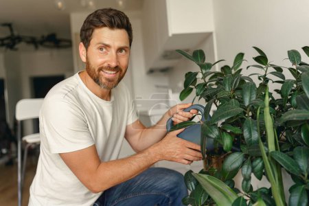 Téléchargez les photos : Homme mûr souriant arrosant des plantes à la maison. Travaux ménagers et concept de ménage. Photo de haute qualité - en image libre de droit