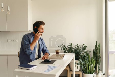 Téléchargez les photos : Homme freelance en vêtements décontractés parler par téléphone tout en travaillant sur ordinateur portable sur la cuisine à la maison - en image libre de droit