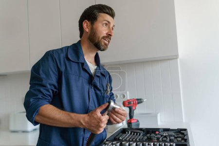 Téléchargez les photos : Travailleur masculin avec boîte à outils debout sur fond de cuisine à la maison et regardant côté. Photo de haute qualité - en image libre de droit