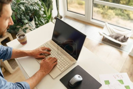 Téléchargez les photos : Homme freelance en vêtements décontractés travaillant sur ordinateur portable sur la cuisine à la maison. Concept de travail à distance - en image libre de droit