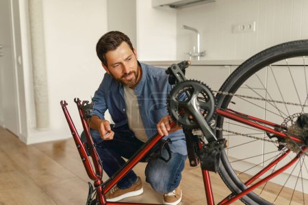 Téléchargez les photos : Beau souriant homme en vêtements décontractés réparer le vélo lui-même à la maison. Photo de haute qualité - en image libre de droit