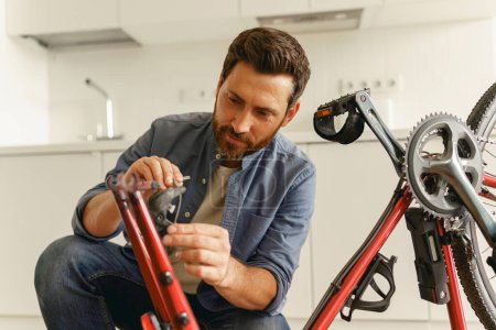 Téléchargez les photos : Beau souriant homme en vêtements décontractés réparer le vélo lui-même à la maison. Photo de haute qualité - en image libre de droit