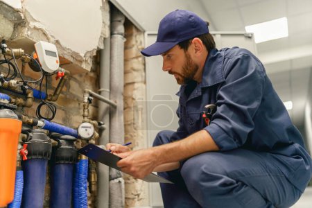 Téléchargez les photos : Handyman vérifie les tuyaux dans le système d'alimentation en eau avant de réparer et prend des notes dans le presse-papiers - en image libre de droit