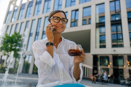 Téléchargez les photos : Femme d'affaires souriante avec tasse de café téléphone parlant assis sur fond de bâtiment moderne - en image libre de droit