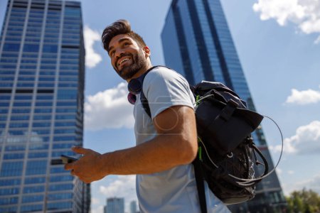 Téléchargez les photos : Homme souriant avec sac à dos et téléphone debout sur fond de gratte-ciel et regarde loin - en image libre de droit