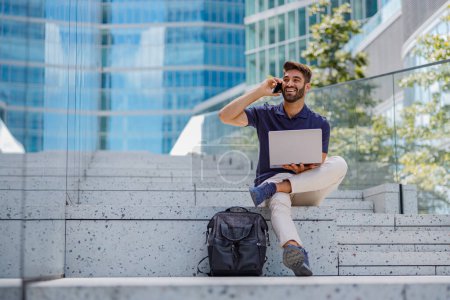 Téléchargez les photos : Directeur des ventes masculins joyeux travaillant sur ordinateur portable et téléphone parlant assis sur fond de gratte-ciel - en image libre de droit