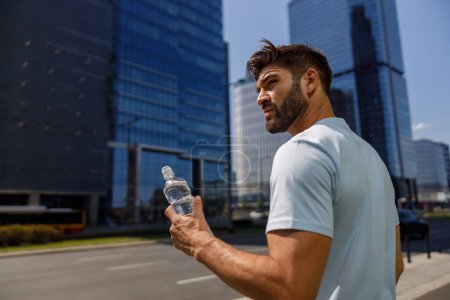 Téléchargez les photos : Bel homme de sport boire de l'eau de la bouteille après avoir couru dans les gratte-ciel de la ville fond - en image libre de droit