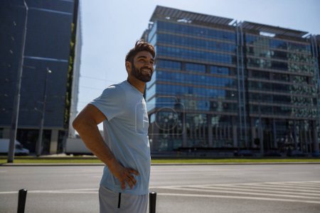 Photo for Sporty man in sportswear resting after training standing on modern city street background - Royalty Free Image