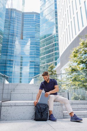 Foto de Sonriente hombre de negocios poniendo portátil dentro de la mochila mientras está sentado en los escalones en el fondo de los rascacielos - Imagen libre de derechos