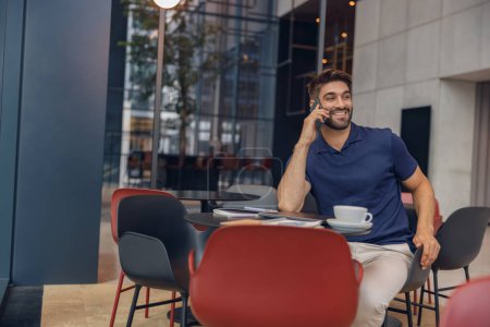 Téléchargez les photos : Beau freelance masculin parlant par téléphone avec le client assis dans un café confortable. Concept de travail à distance - en image libre de droit